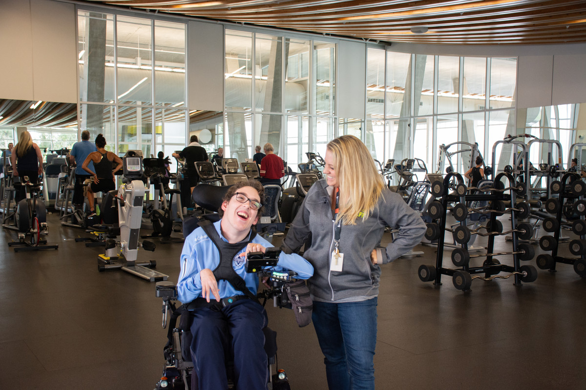 Nathan, a power wheelchair user is all smiles with city staff member at the accessible gym full of adaptive equipment. 