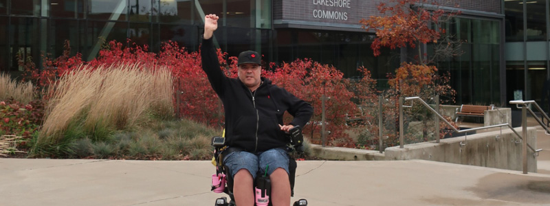 Man in a wheelchair holding on arm in the air. Wearing a black hat and sweater with shorts.