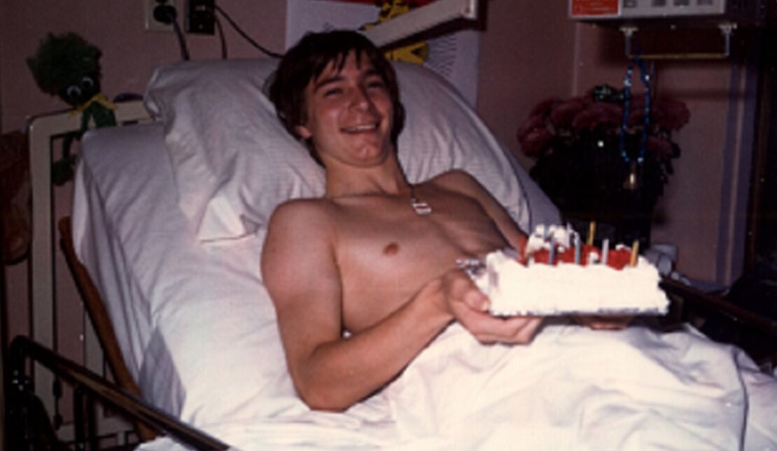 Rick Hansen smiles as he lays on hospital bed with cake during his 16th birthday