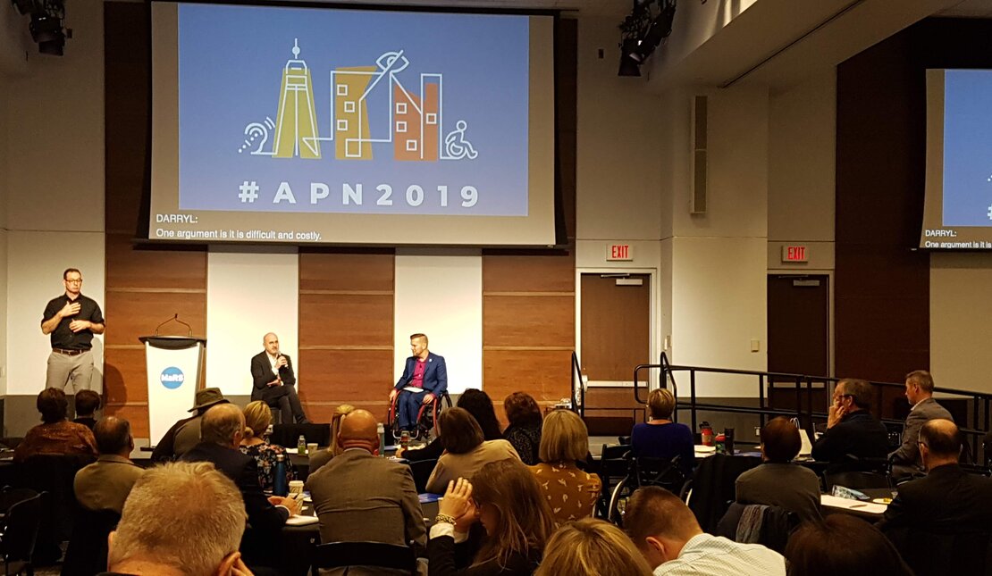 APN 2019. A conference room filled with people looking at two speakers at the front of the room. One of the speakers is using a wheelchair. There is an ASL interpreter and a projector that says #APN2019