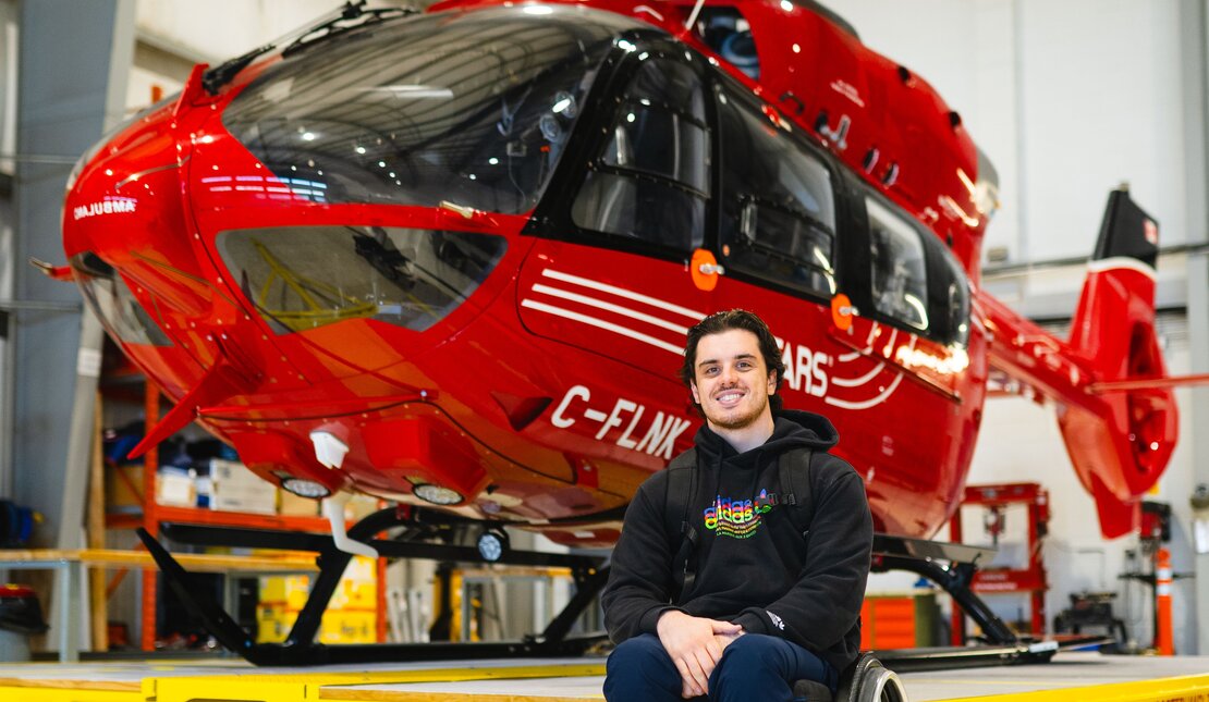 Ryan in front of a large red helicopter inside a garage. Ryan is using a wheelchair.