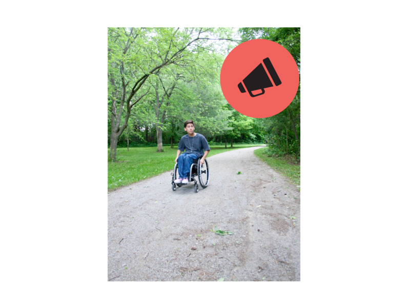 Teenager in wheelchair wheeling down a paved pathway in the middle of a park.