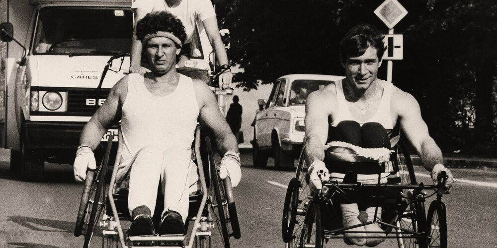 Rick Hansen wheeling in Czechoslovakia with another wheelchair athlete, accompanied by Amanda Reid following behind on a bicycle.