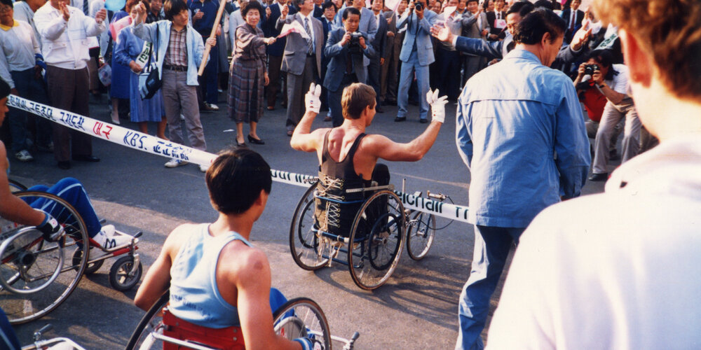 Rick Hansen going through a banner with Korean Broadcasting KBS on it