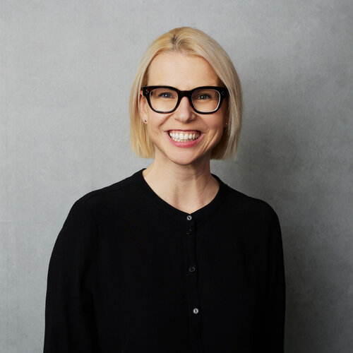 Sarah McCarthy who has chin-length blonde hair and is wearing thick black glasses and a black top. She is in front of a grey wall.