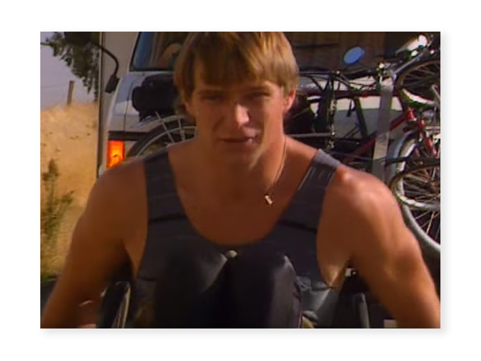 Screenshot from "Power of the Dream" video - a close up of Rick Hansen wheeling during the Man In Motion World Tour, wearing a black shirt and pants. Behind him the front of the Tour motorhome can be seen.
