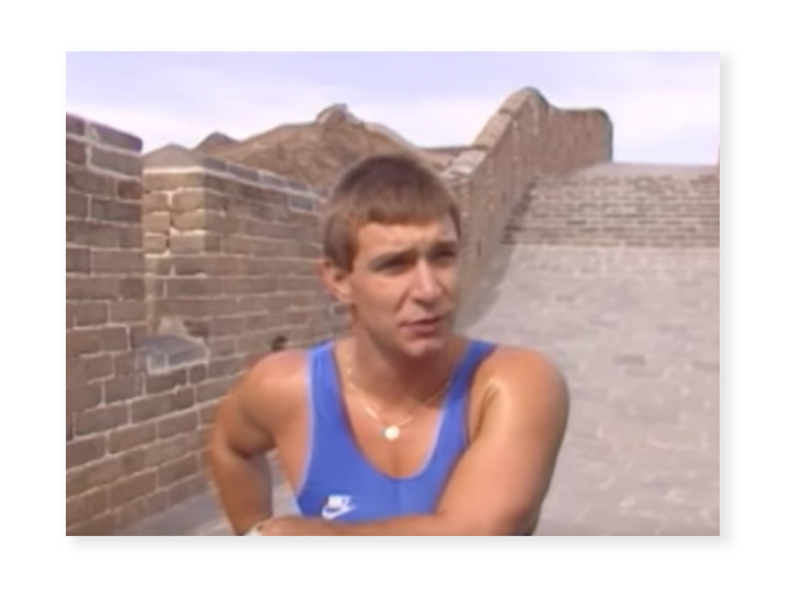 Screenshot from "Rick Hansen Man In Motion World Tour - Great Wall of China" video of Rick Hansen from his chest up on the Great Wall of China. He is wearing a blue track shirt with a white Nike Logo, and looks to be answering a question.