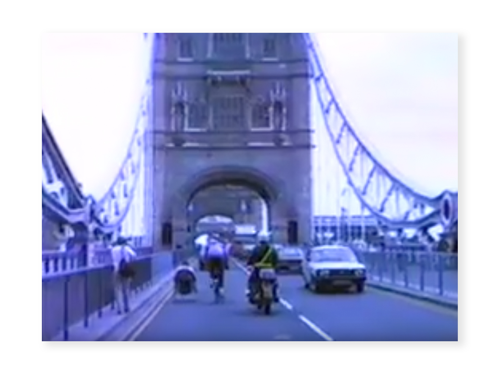 Screenshot from "Rick Hansen Man In Motion World Tour Footage - NA & Europe" video - a blurry image of Rick Hansen wheeling, a Tour team member cycling, and a police escort on a motorbike across London Bridge.