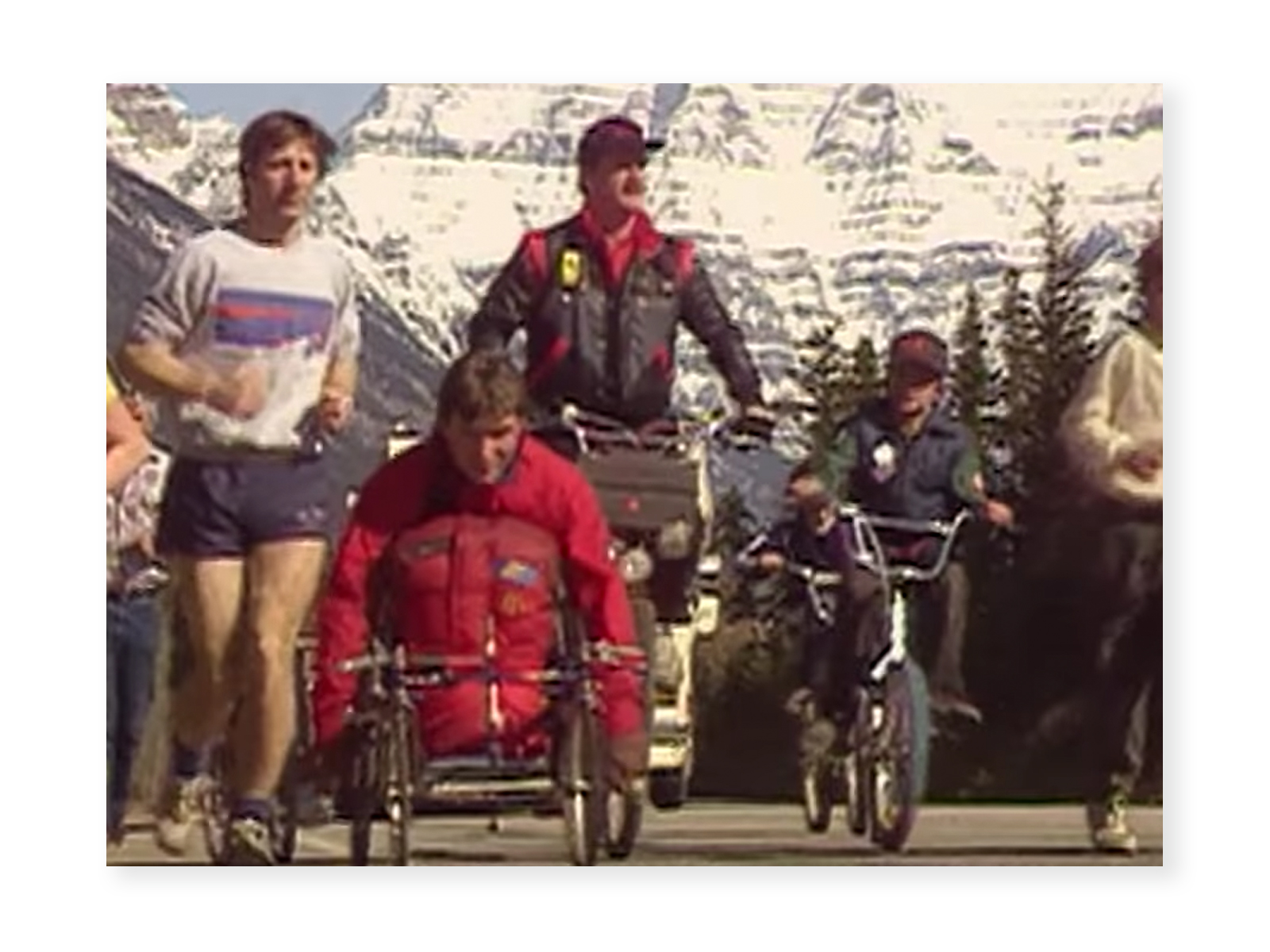 Screenshot from "The Journey" video - an image from the Man In Motion World Tour, where Rick is wheeling wearing a red snow suit. Beside him are a few runners as well as cyclists. In the background is a mountain with snow and evergreen trees.