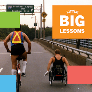 View from behind Rick Hansen as he wheels along a US highway accompanied by a cyclist on his left. Little Big Lessons logo text.