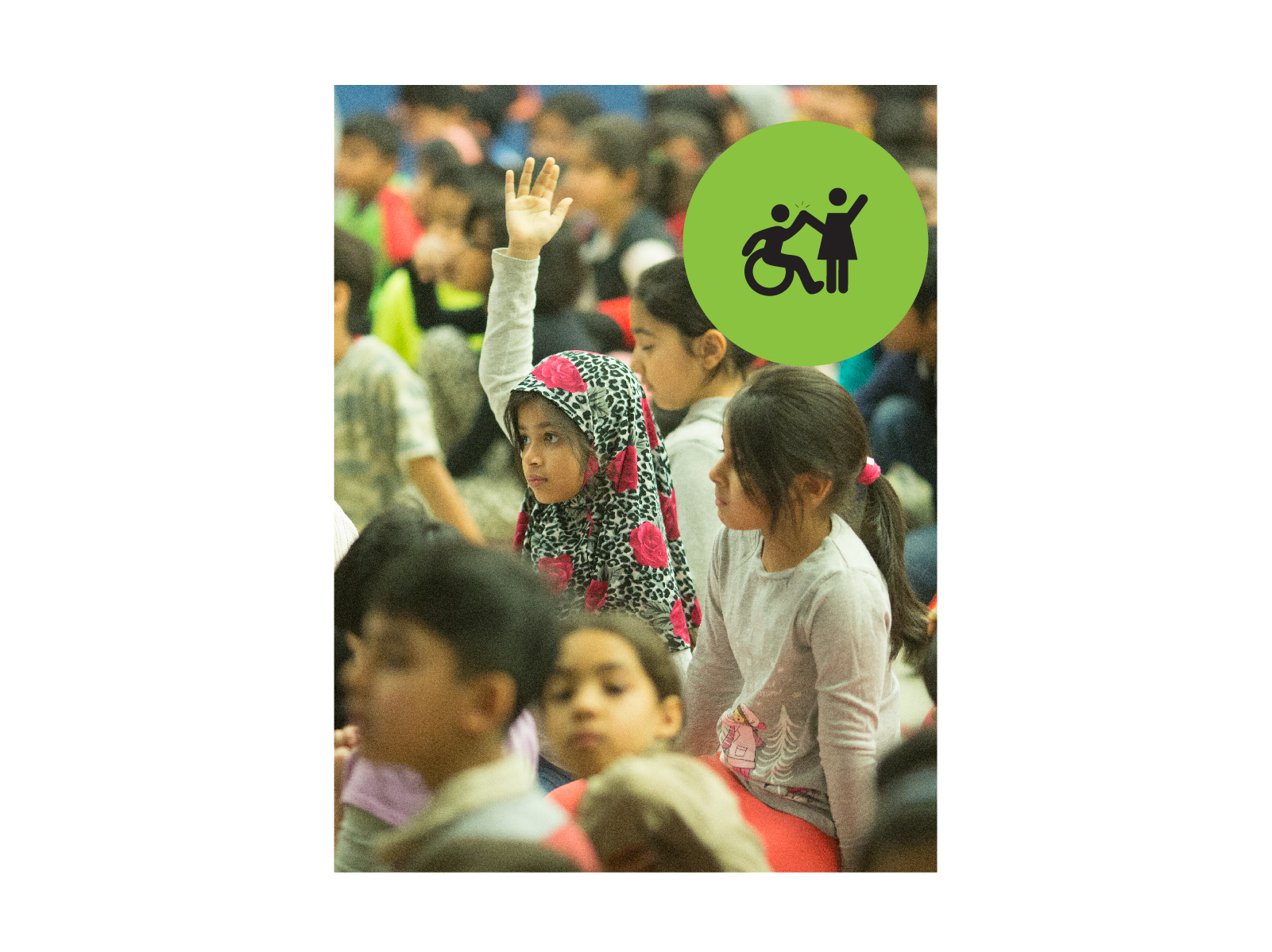 A girl raising her hand in a crowded assembly. Small green circle icon with graphic of person in a wheelchair high-fiving a person that is standing.