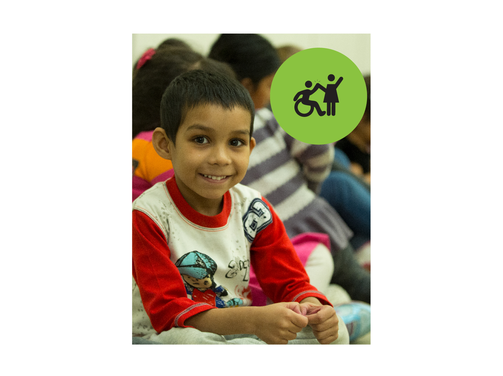 Young boy wearing a red and white long-sleeved shirt sitting and smiling at camera. Small green circle icon with graphic of person in a wheelchair high-fiving a person that is standing.