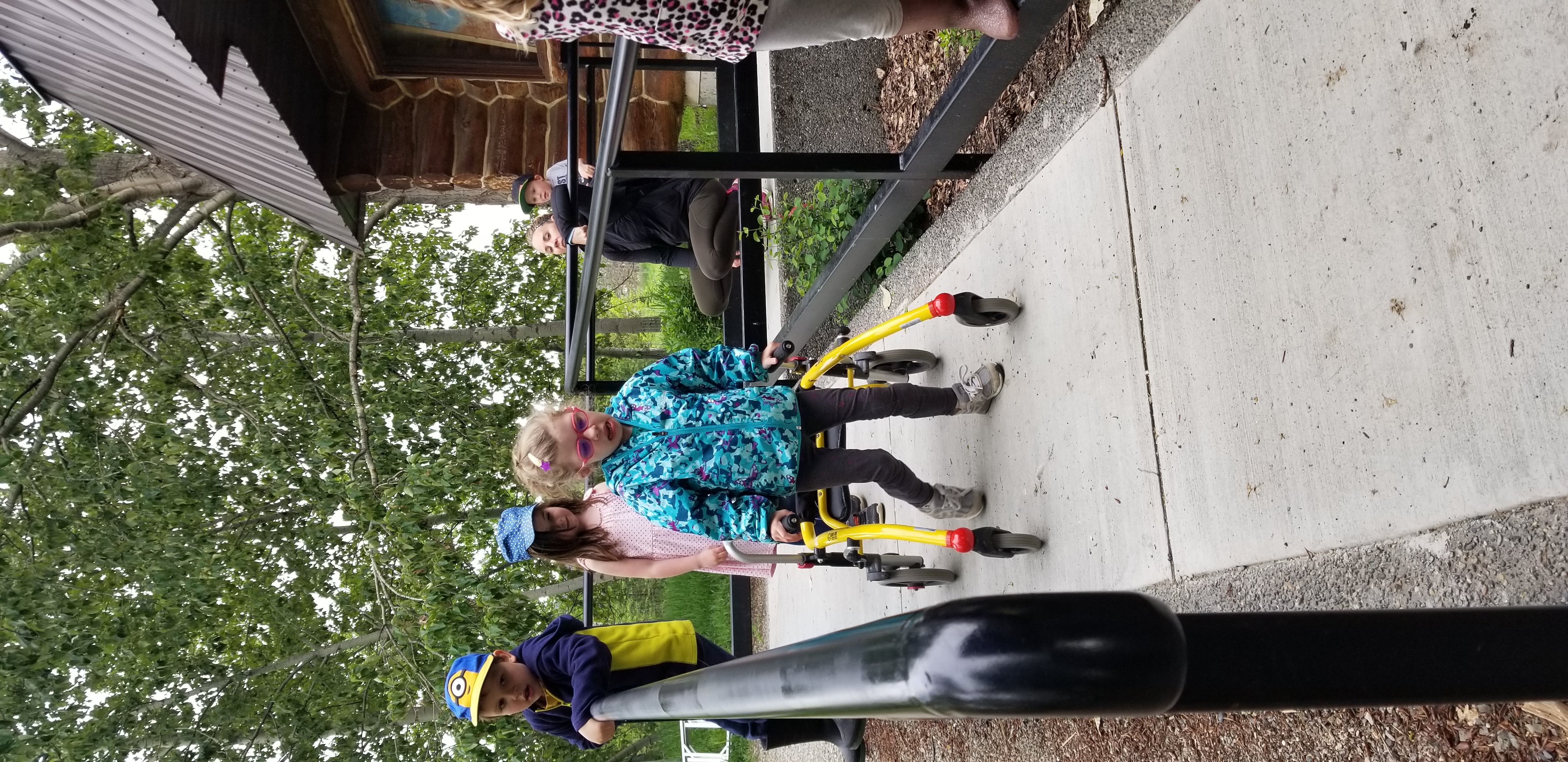 A girl with blonde hair, pink framed glasses, and wearing turquoise jacket and black pants pushes a trolley down the accessible ramp outside Scout Island Nature Centre. Behind her are two children, a girl and a boy following her down while a teach and another girl watch.