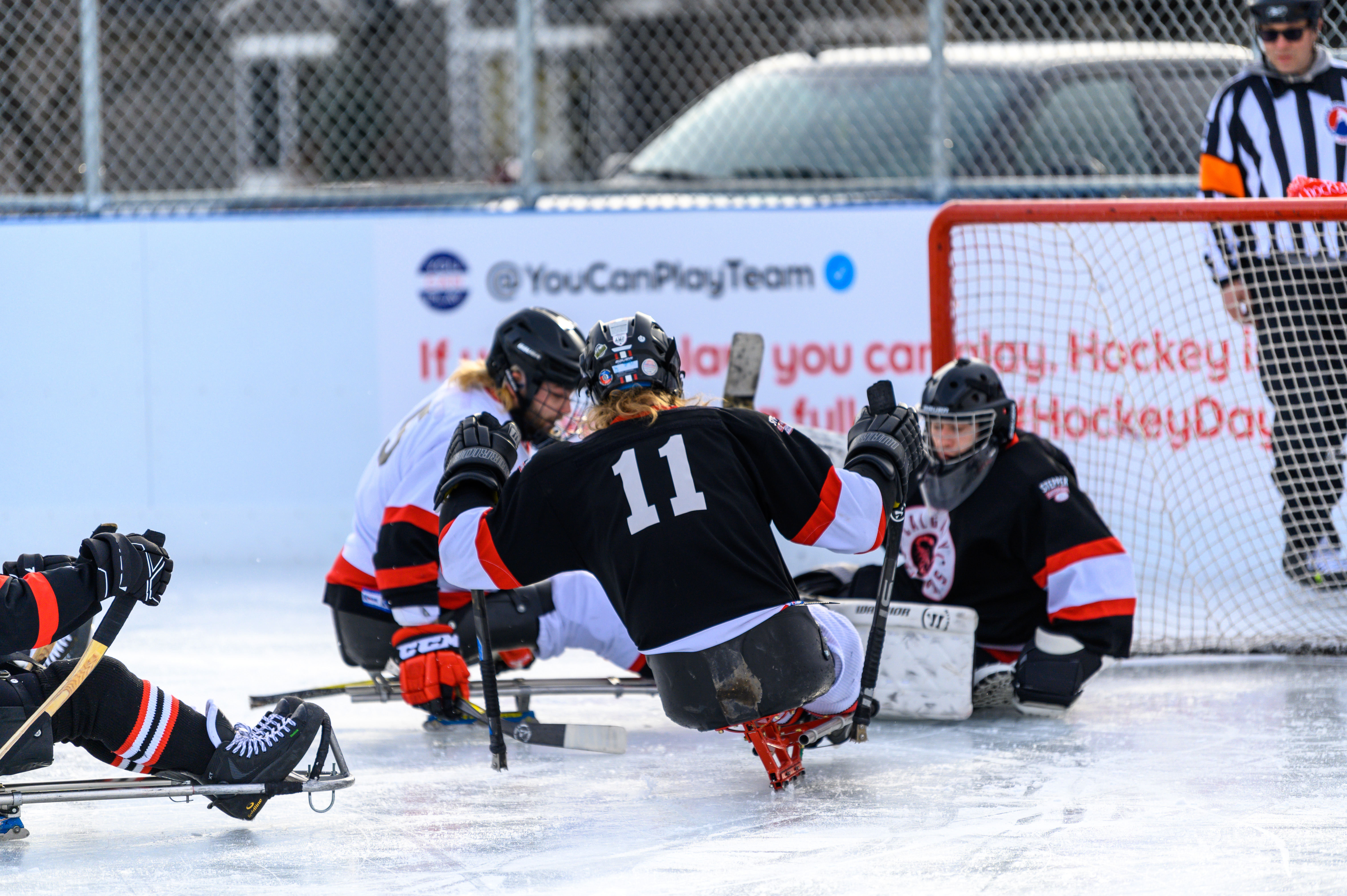 People playing sledge hockey close to the net. One team is wearing black jerseys and the other team is wearing white jerseys.