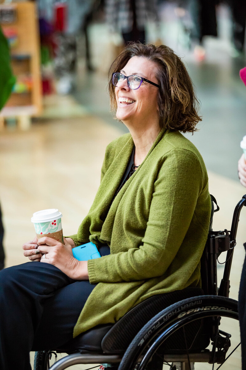 Stephanie Cadieux, who has shoulder length brown hair and glasses. She is wearing a green cardigan with dark coloured pants and holding a Starbucks cup. She is using a wheelchair and is smiling away from the camera.