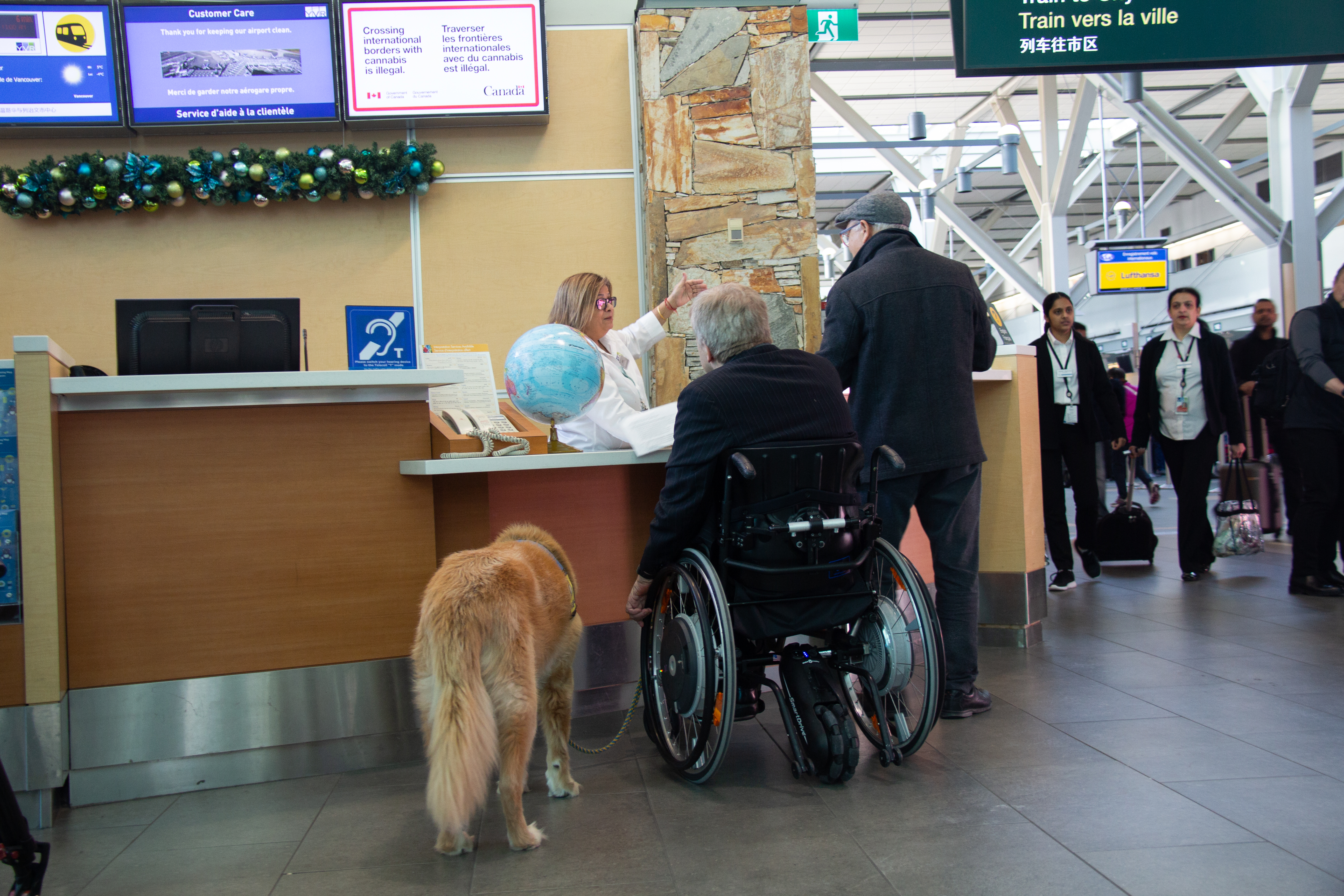 Two older men are asking for information at an airport reception desk. One of the men is in a wheelchair and has a service dog. There is a blue sign on the reception desk with a white icon that indicates the building is accessible for those with hearing loss.