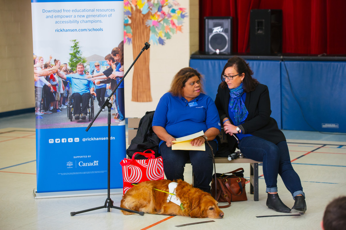 Two woman lean together to speak with a guide dog at their feet asleep