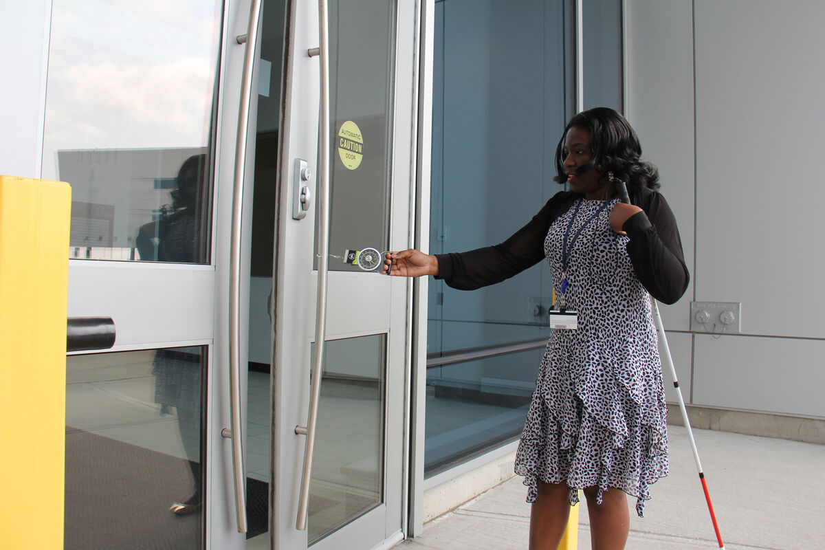 Woman with shoulder-length dark hair standing outside a glass entrance, using a white cane while reaching toward an automatic door button.