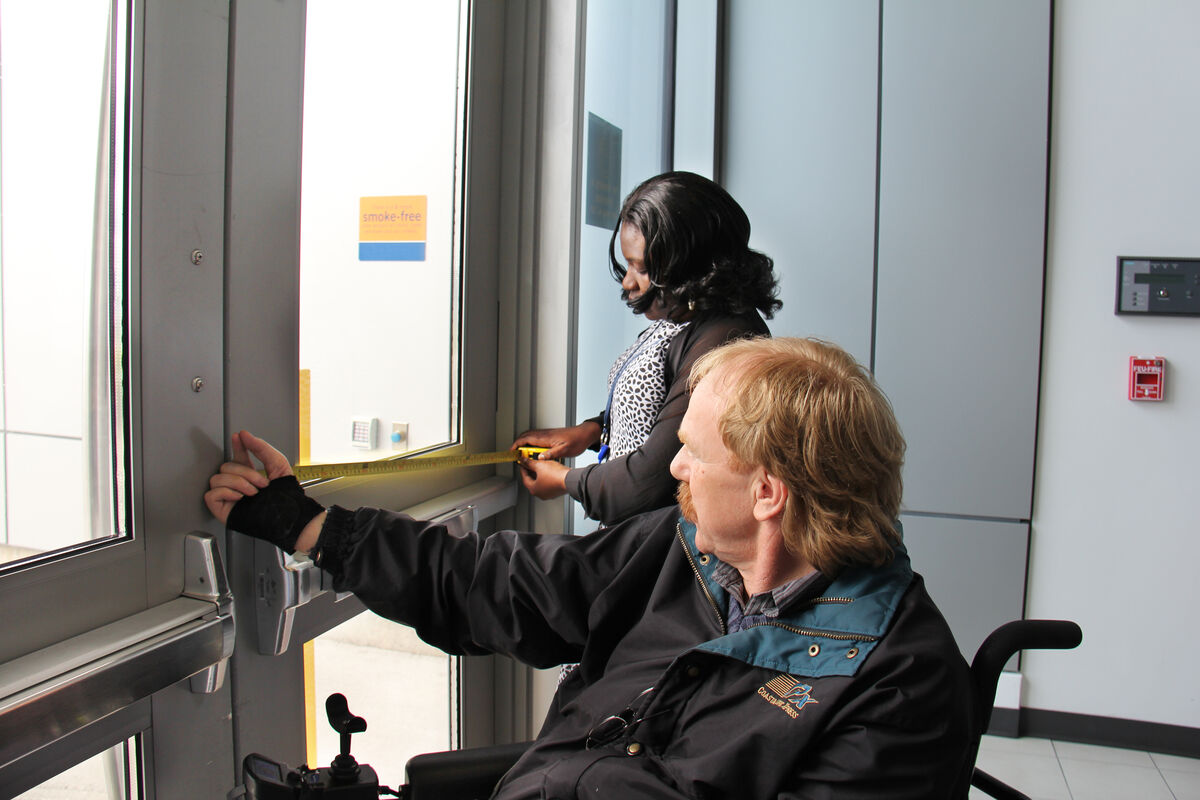 Person using a a power wheelchair holds a door open while another person measures the width of the doorway with a tape measure.