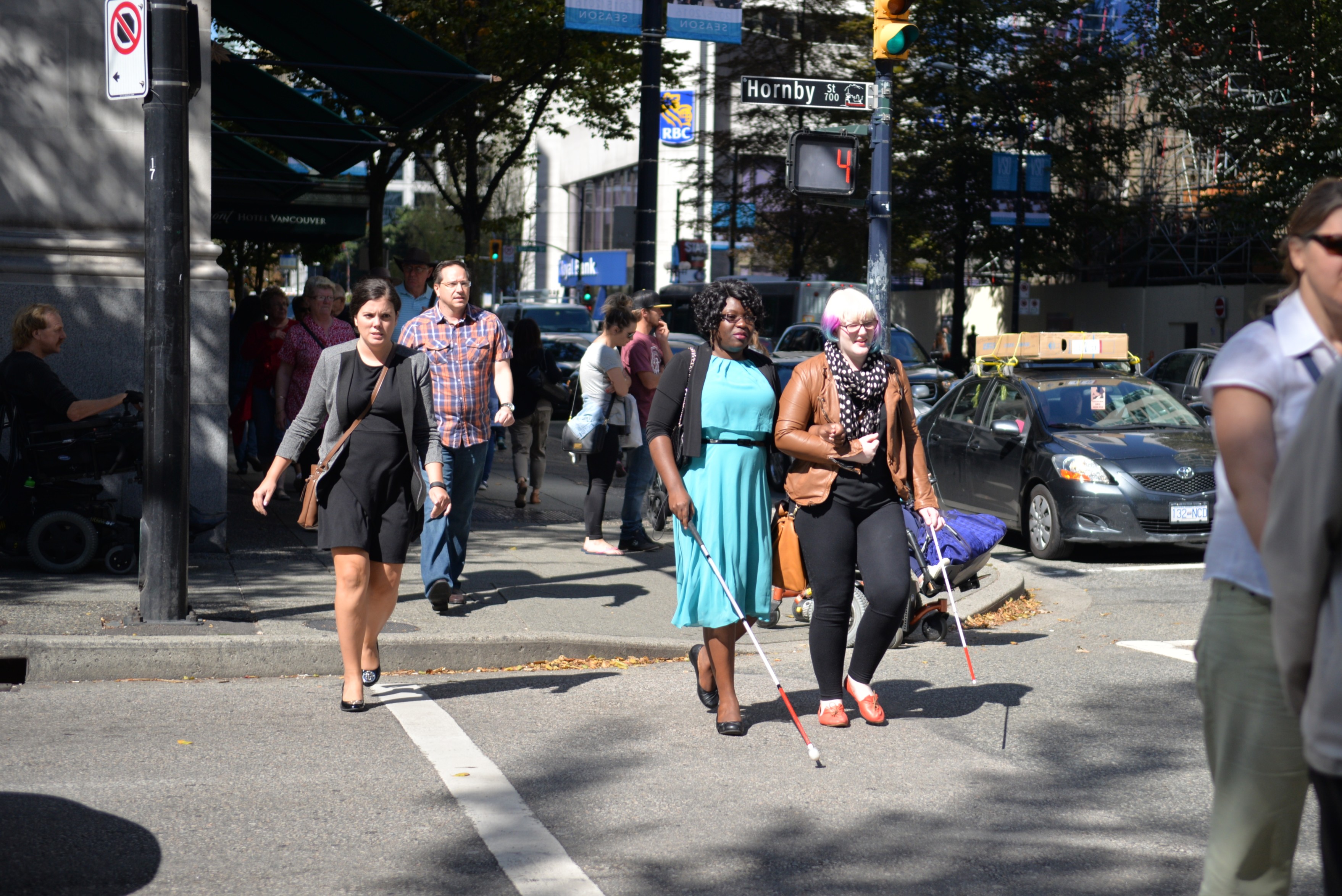 A busy city crosswalk with multiple pedestrians walking across the street. Two individuals in the foreground are using white canes for navigation