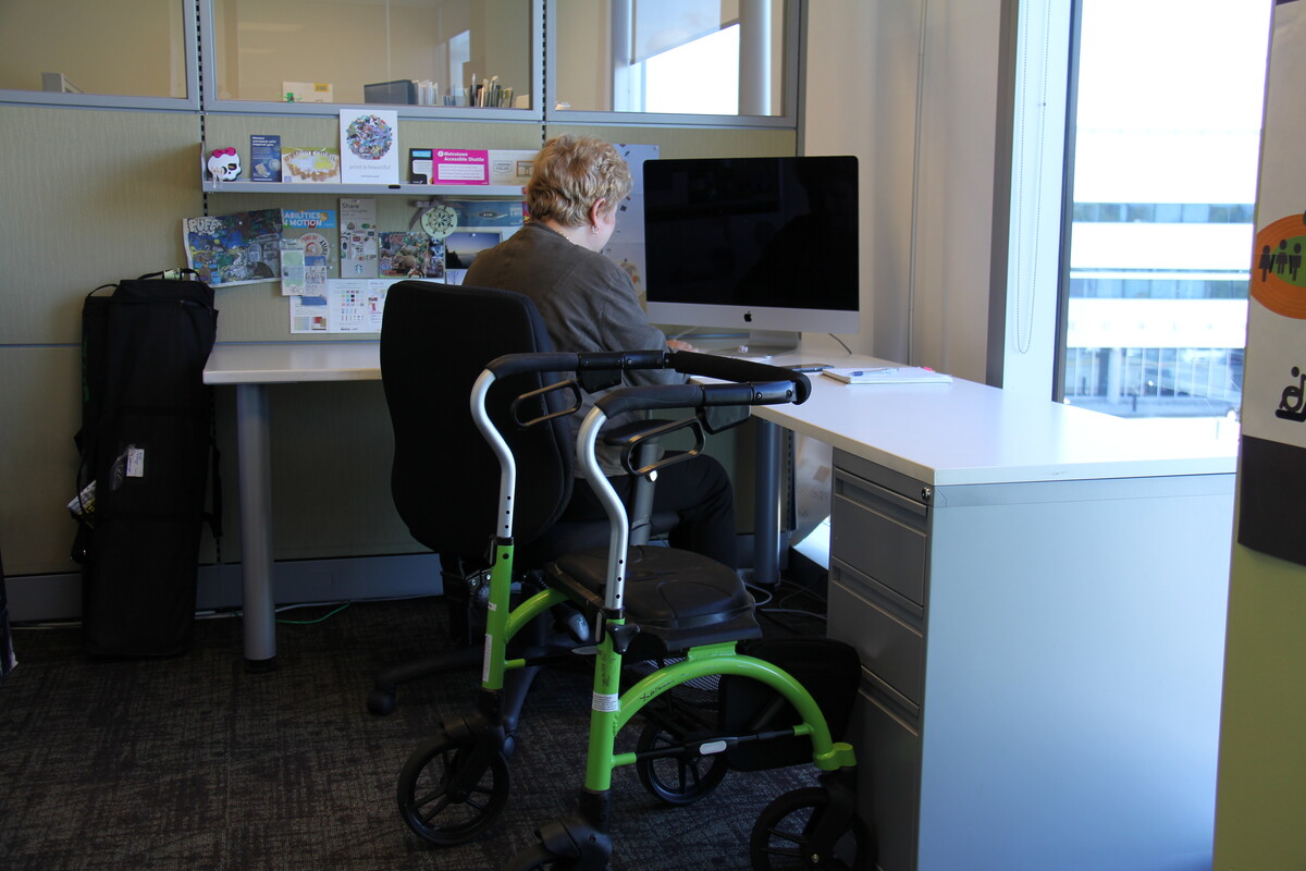 A person with short blonde hair using a computer at a desk. There is an assistive walking device beside the desk.