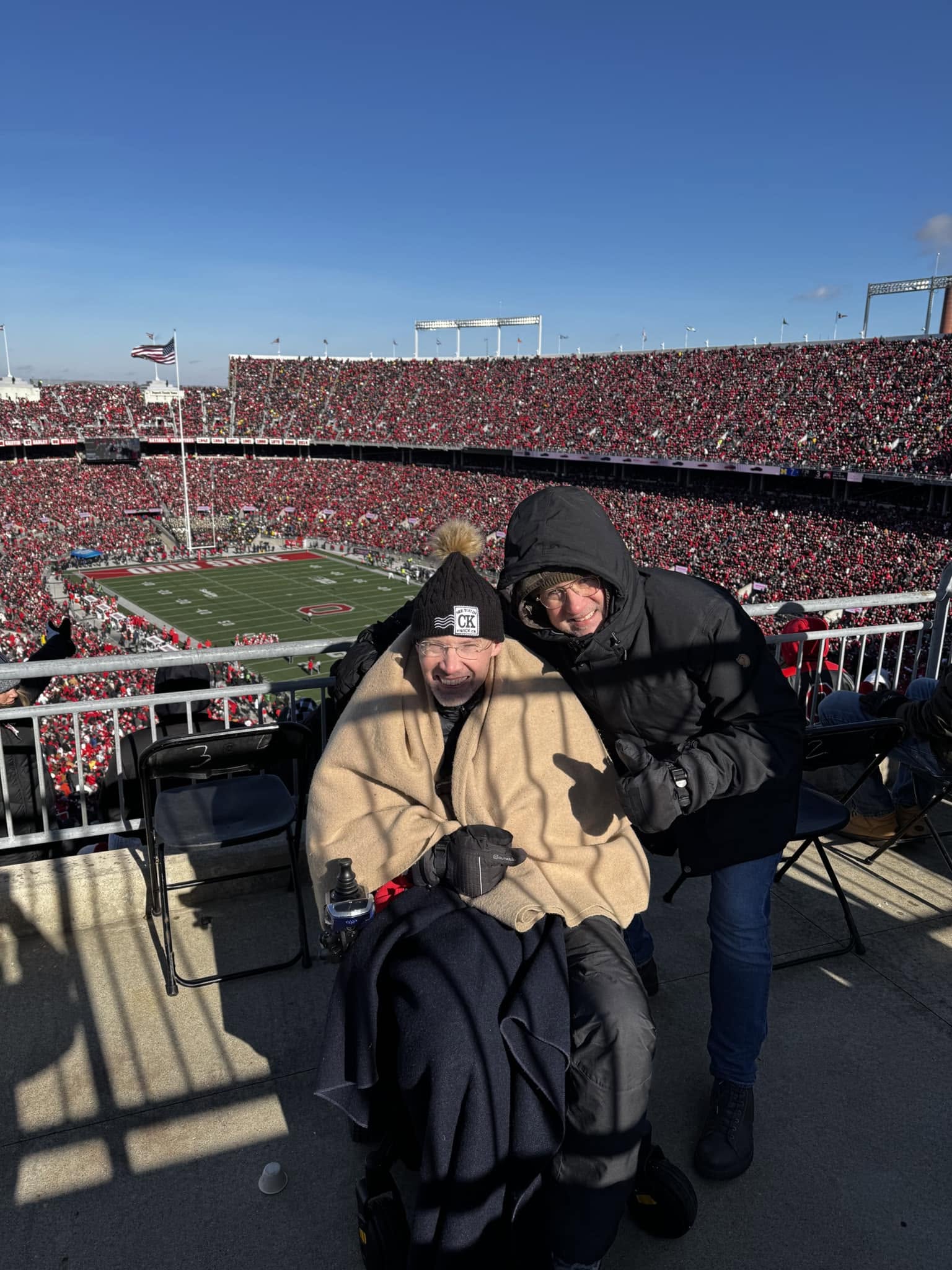 Two people bundled in winter clothing pose in the stands of a packed football stadium. One person is seated in a power wheelchair and wrapped in a tan blanket, with the football field and crowd in the background.