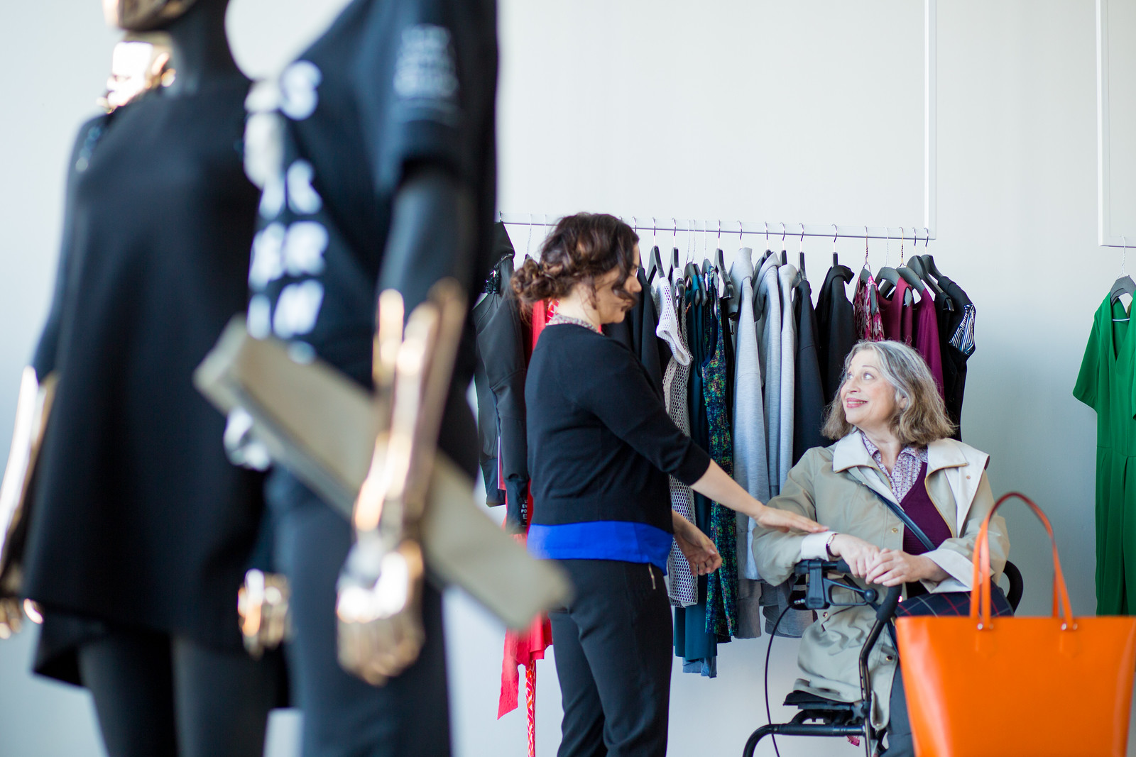 An elderly woman shops for a dress while using a wheeled mobility device. A saleswoman holds up a dress.