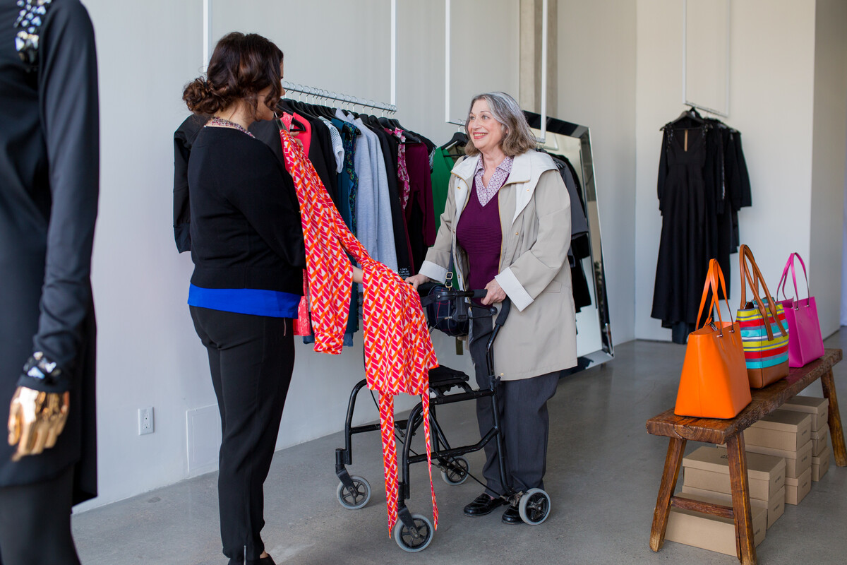 An older woman who has a walker shopping and holding up a clothing item. A younger woman is assisting her.