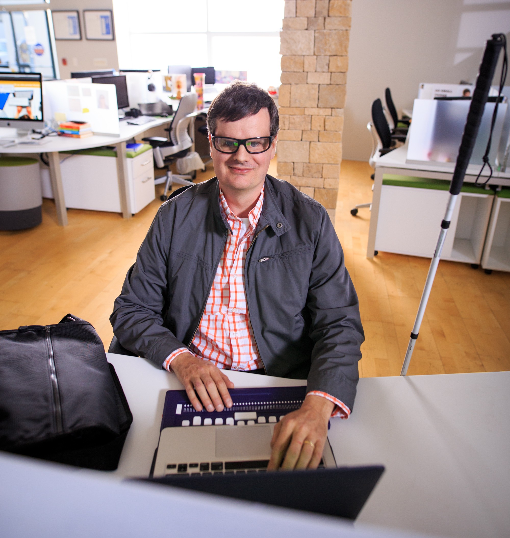 A man sits at a desk in an office, using an accessible Braille keyboard. He faces a computer monitor, with a white cane resting