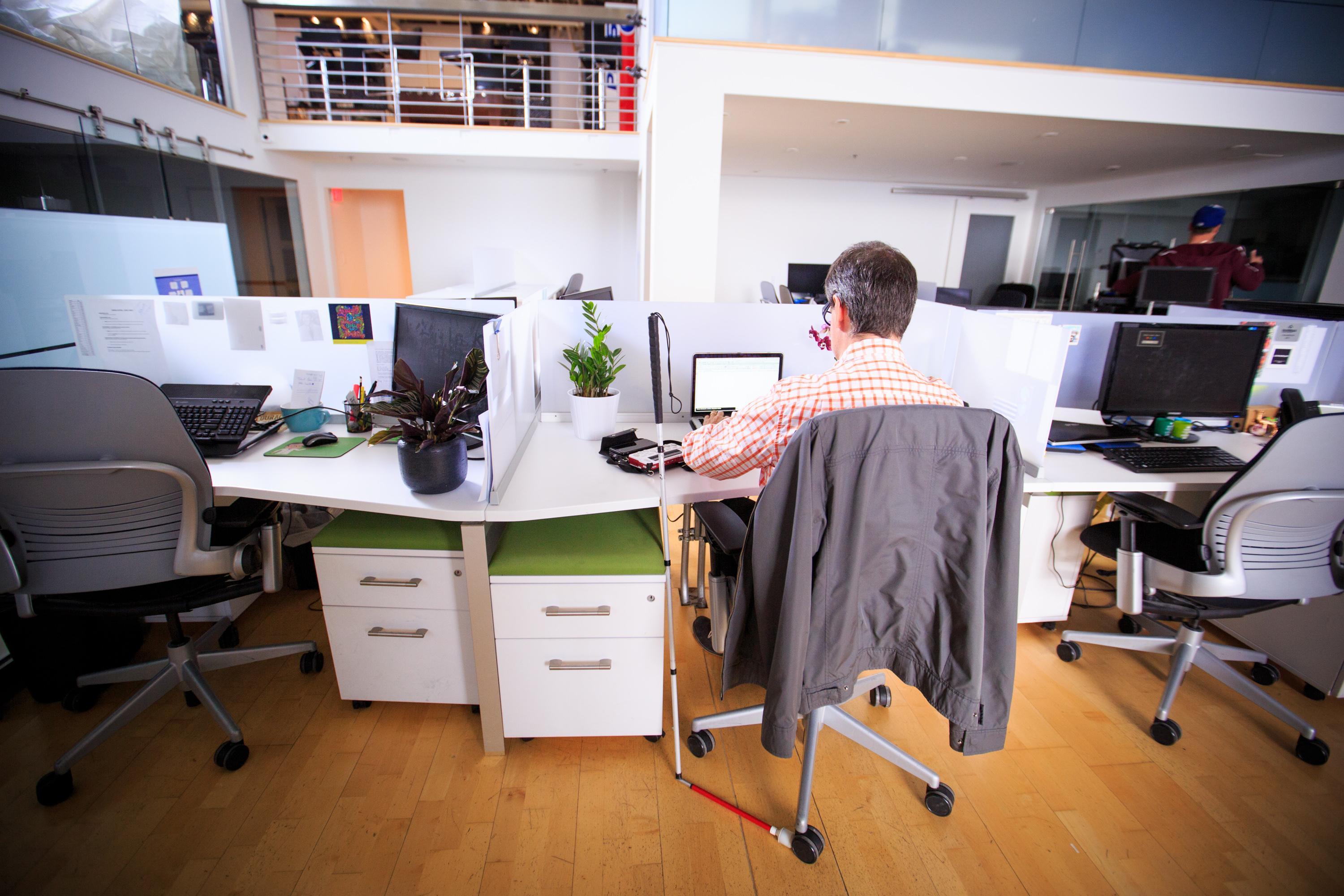 In an office a man sits at the center desk facing a laptop. The background shows glass walls and an upper mezzanine with railing and a white cane on his side