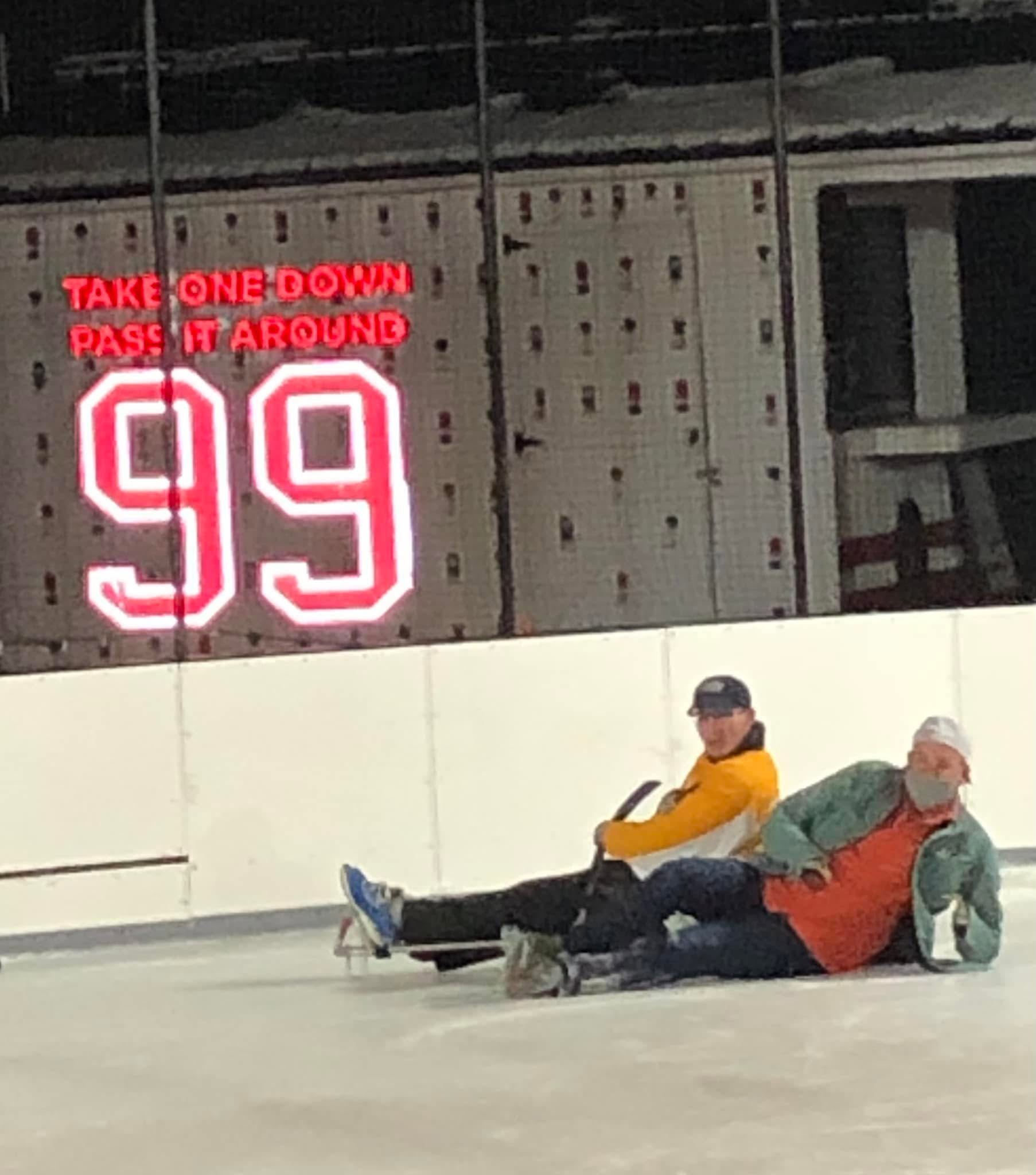 Two people seated on the ice of a hockey rink beneath an illuminated sign reading, “Take One Down Pass It Around 99.”