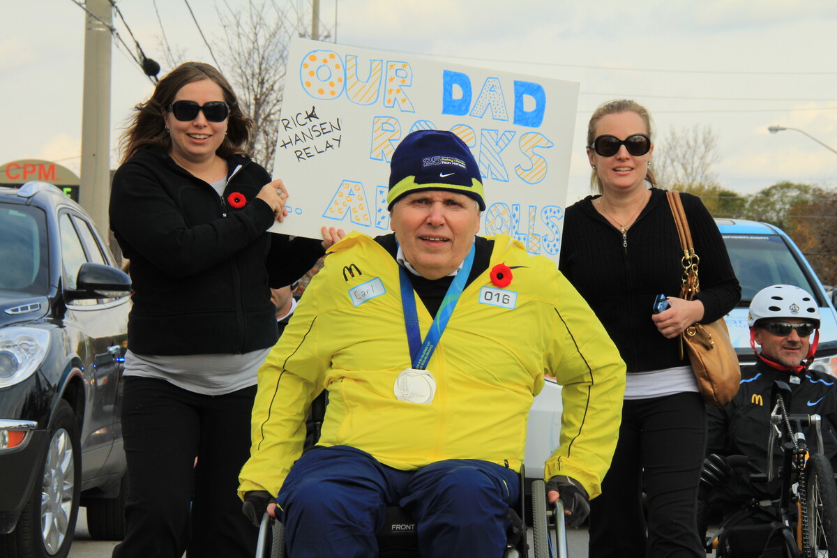 Group participating in a road relay, including a man using a wheelchair wearing a medal and two women walking behind. One participant holds a sign reading “Our Dad Rocks"