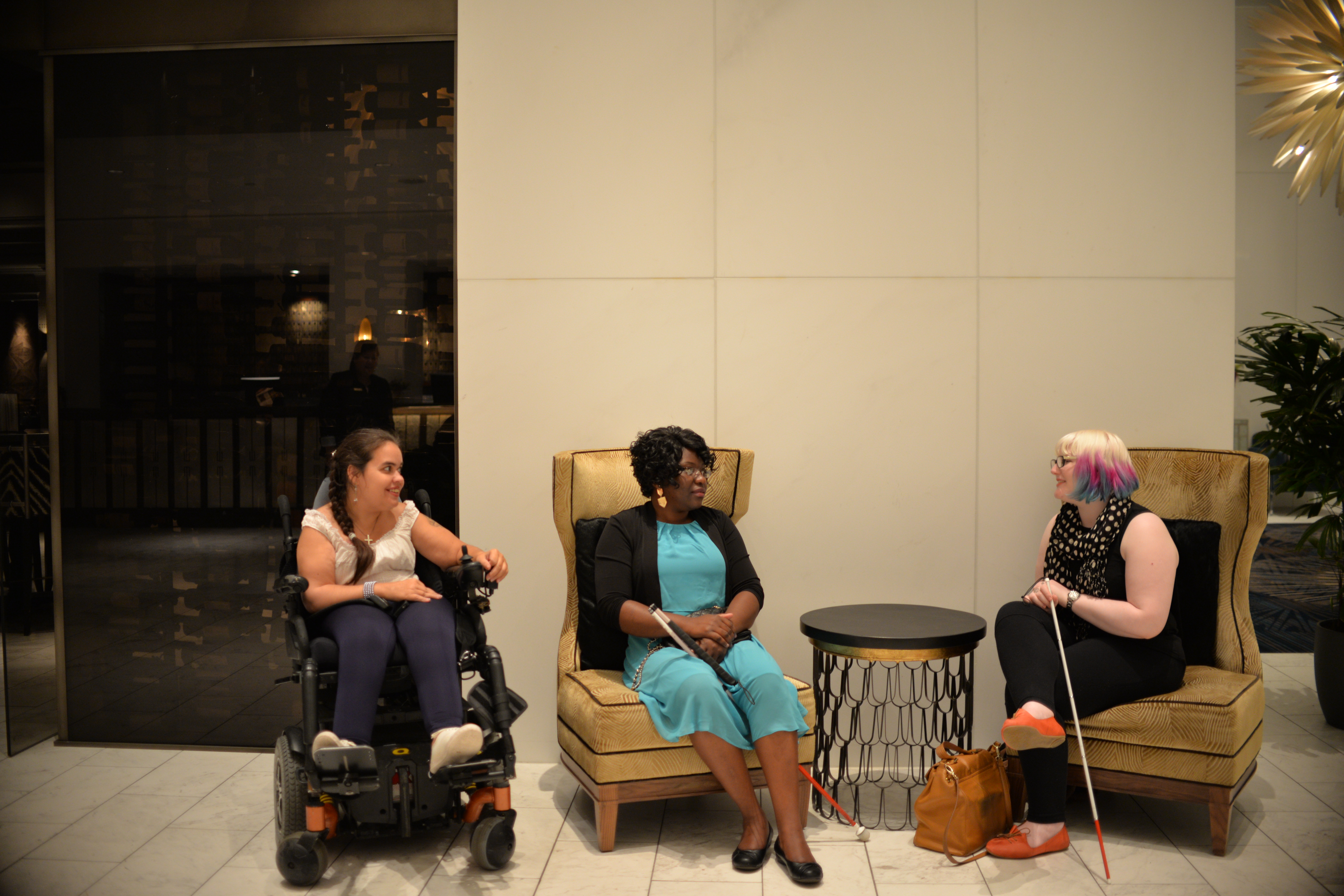 Three people seated in lounge chairs in a modern indoor space, engaged in conversation. One person uses a power wheelchair and another holds a white cane, with a small table between them.