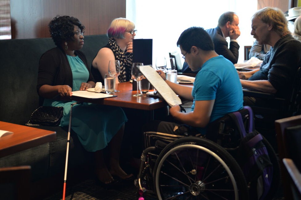 Group seated around a restaurant table. One person uses a wheelchair while reading a menu, and another has a white cane positioned beside the table.