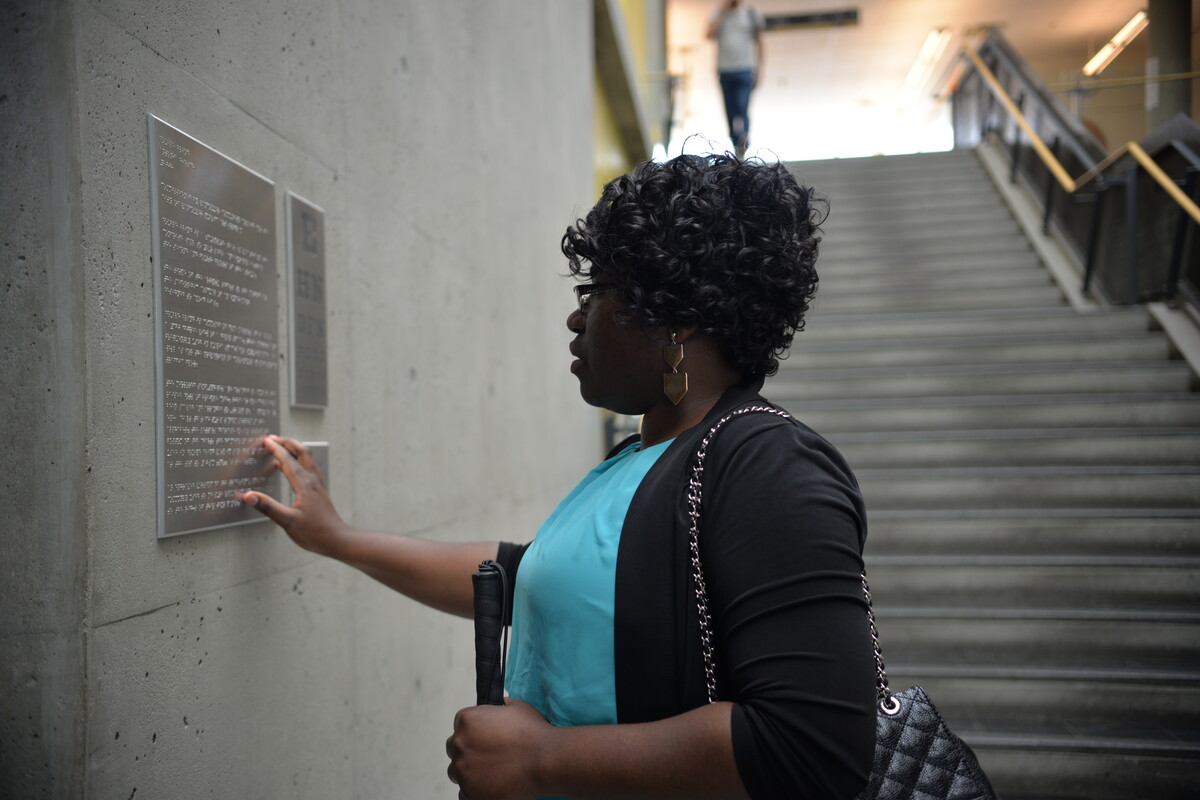 Woman with short dark, curly hair wearing a teal shirt and holding a cane reading braille from a metal sign in a building.