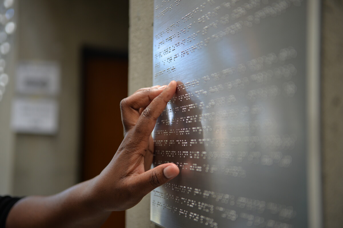 Fingers reading braille on a metal sign that is mounted onto a wall. 