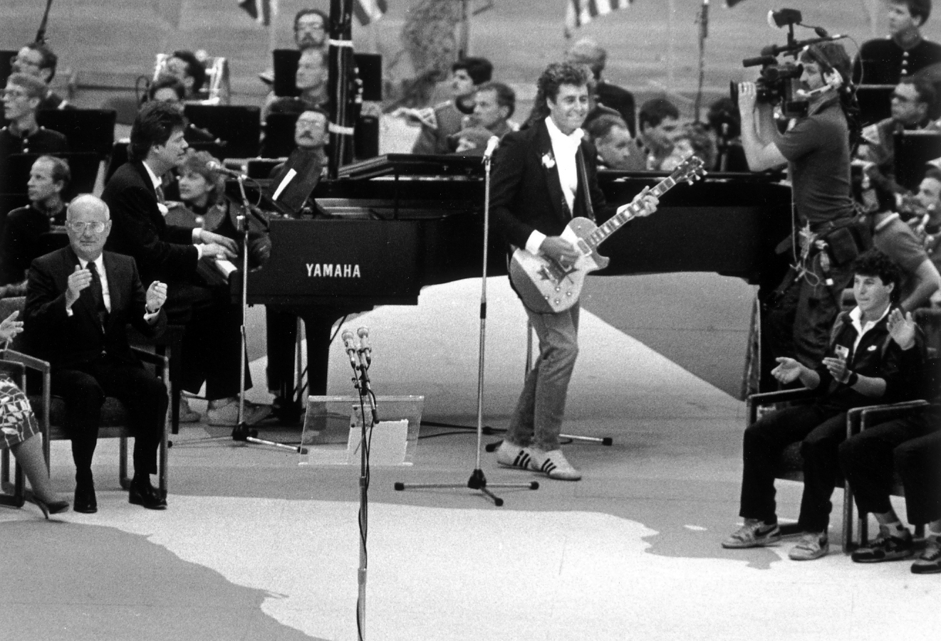 A live musical performance on stage. A man stands playing an electric guitar and wearing a tuxedo jacket with casual sneakers with an orchestra seated further back while several people sit and clap.