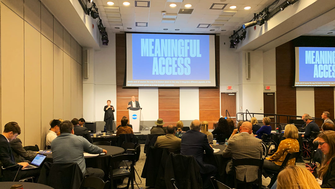 Conference room with attendees seated at round tables facing a speaker at a podium, with a large screen displaying the words “Meaningful Access” and a sign language interpreter beside the stage.