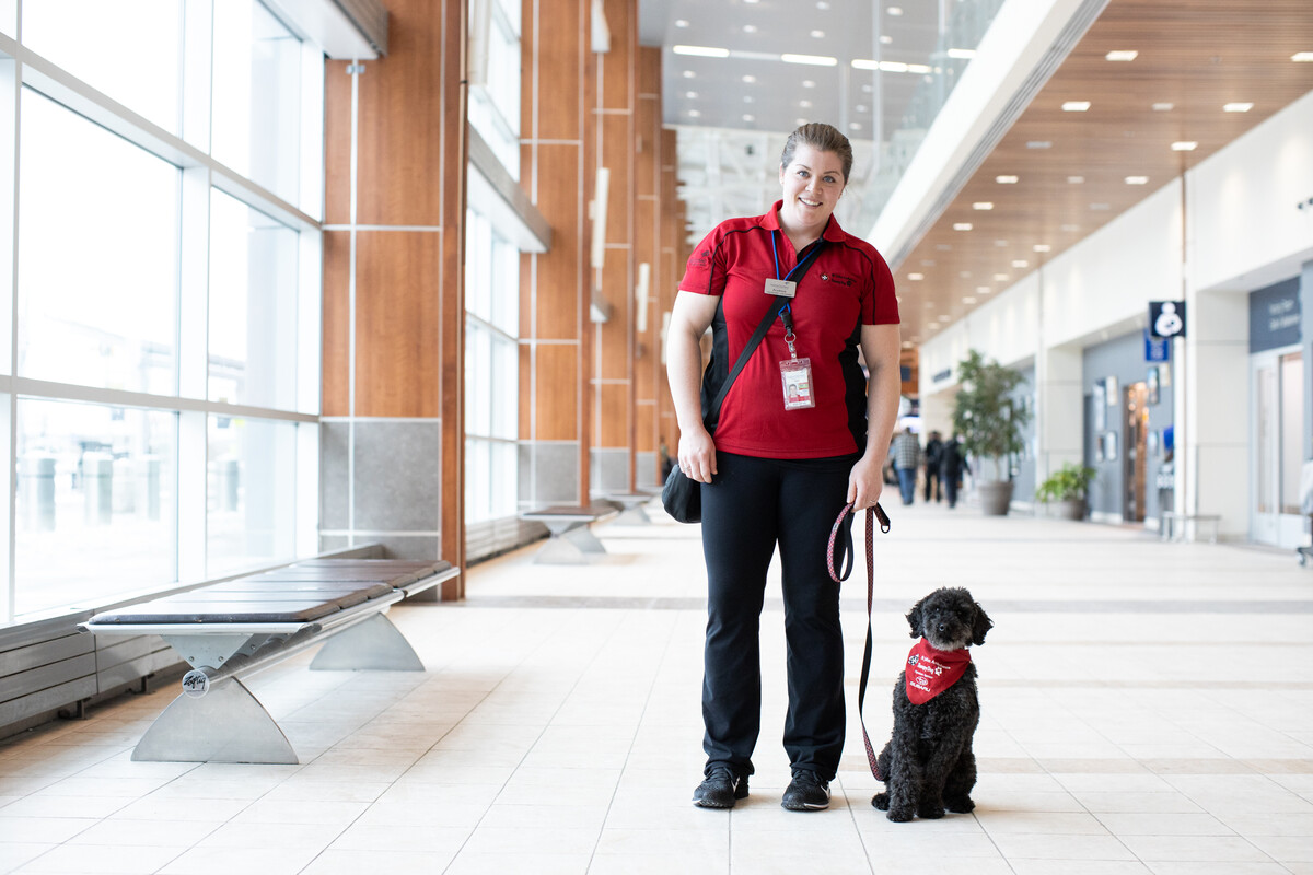Person standing indoors in an airport holding a leash, with a small service dog wearing a red vest seated beside them.