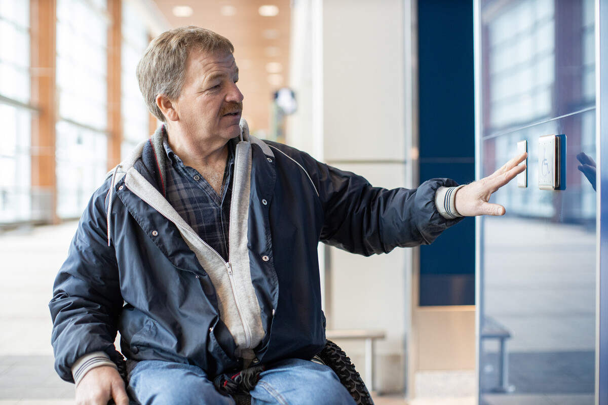 Person using a wheelchair reaches out to touch an accessible wall-mounted control inside a well-lit public building.