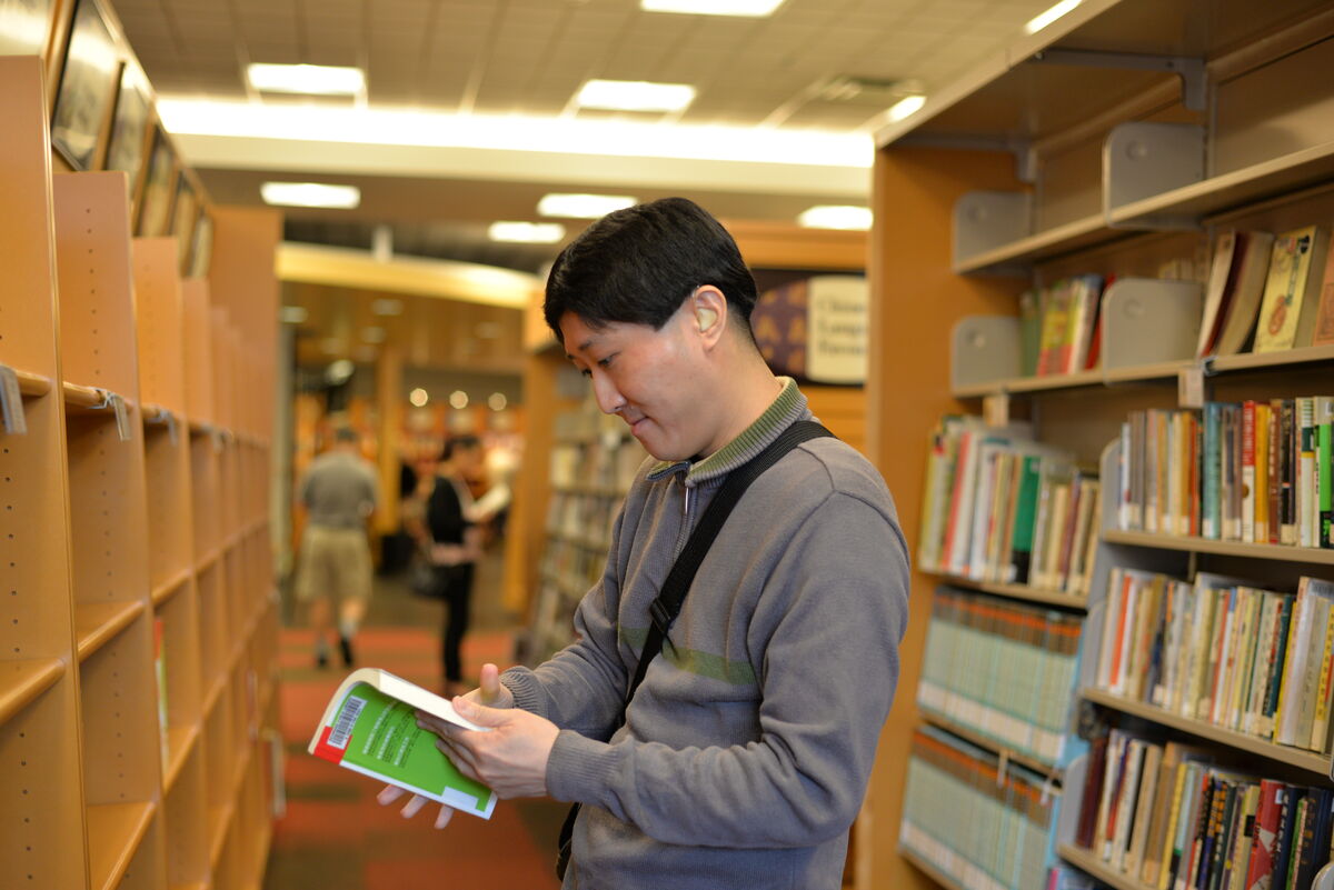Man standing in a library aisle reading a book, with shelves of books on both sides and wearing a hearing aid