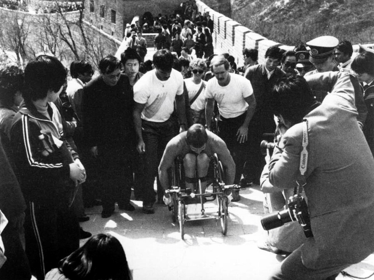 Black and white photo of Rick Hansen surrounded by a crown of people as he pushes up the Great Wall of China.