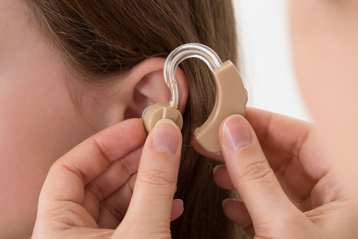 Close-up of a hand placing a beige behind-the-ear hearing aid onto a person’s ear, with brown hair visible around the ear.