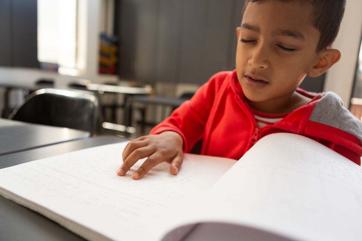 Young child with short dark hair wearing a red sweater sits at a desk reading a Braille book, using one hand to follow the raised dots on the page.