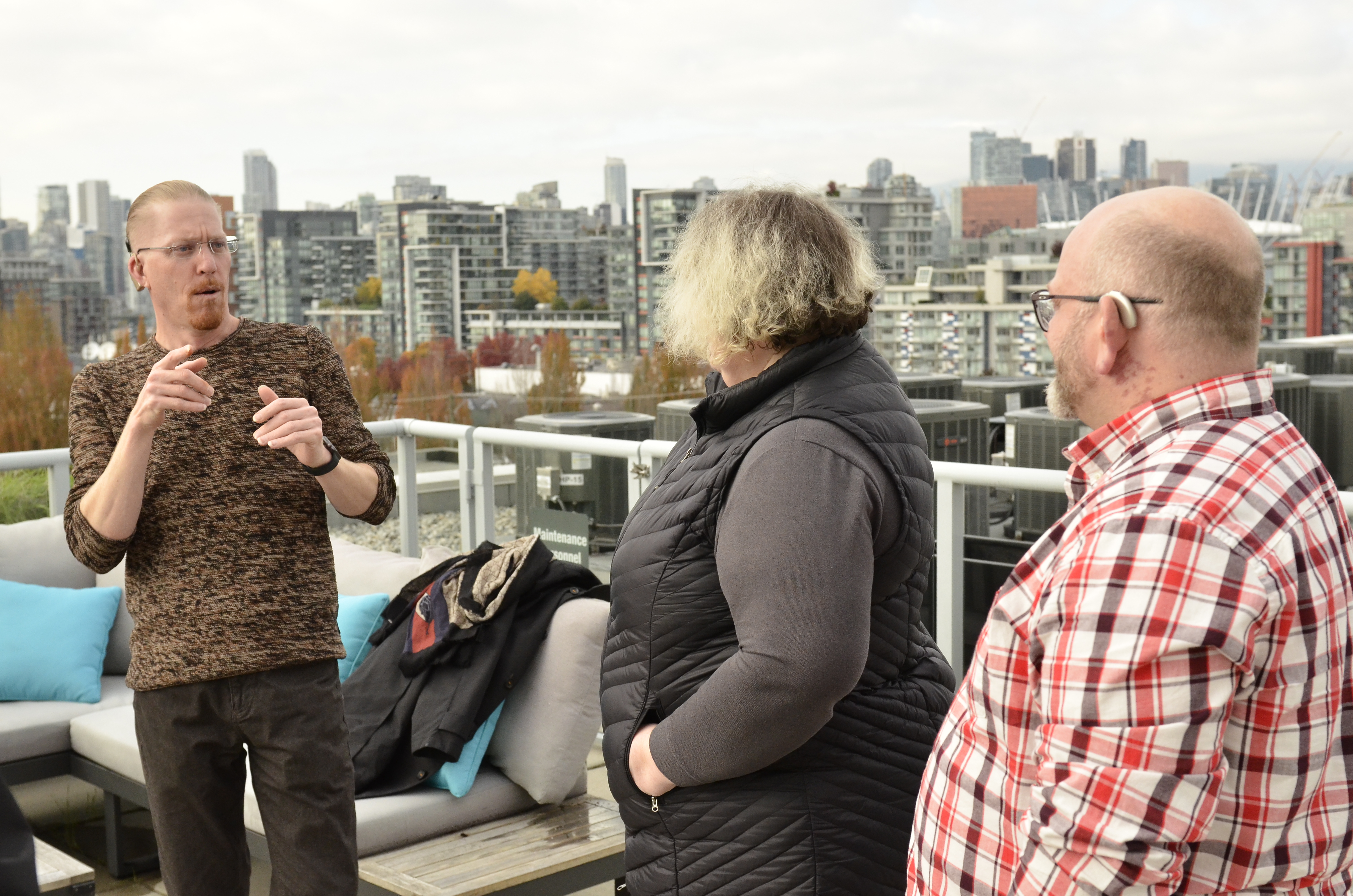 Three people stand on a rooftop terrace. The person on the left gestures with both hands, while the other two face them – one of whom is wearing a hearing aid.