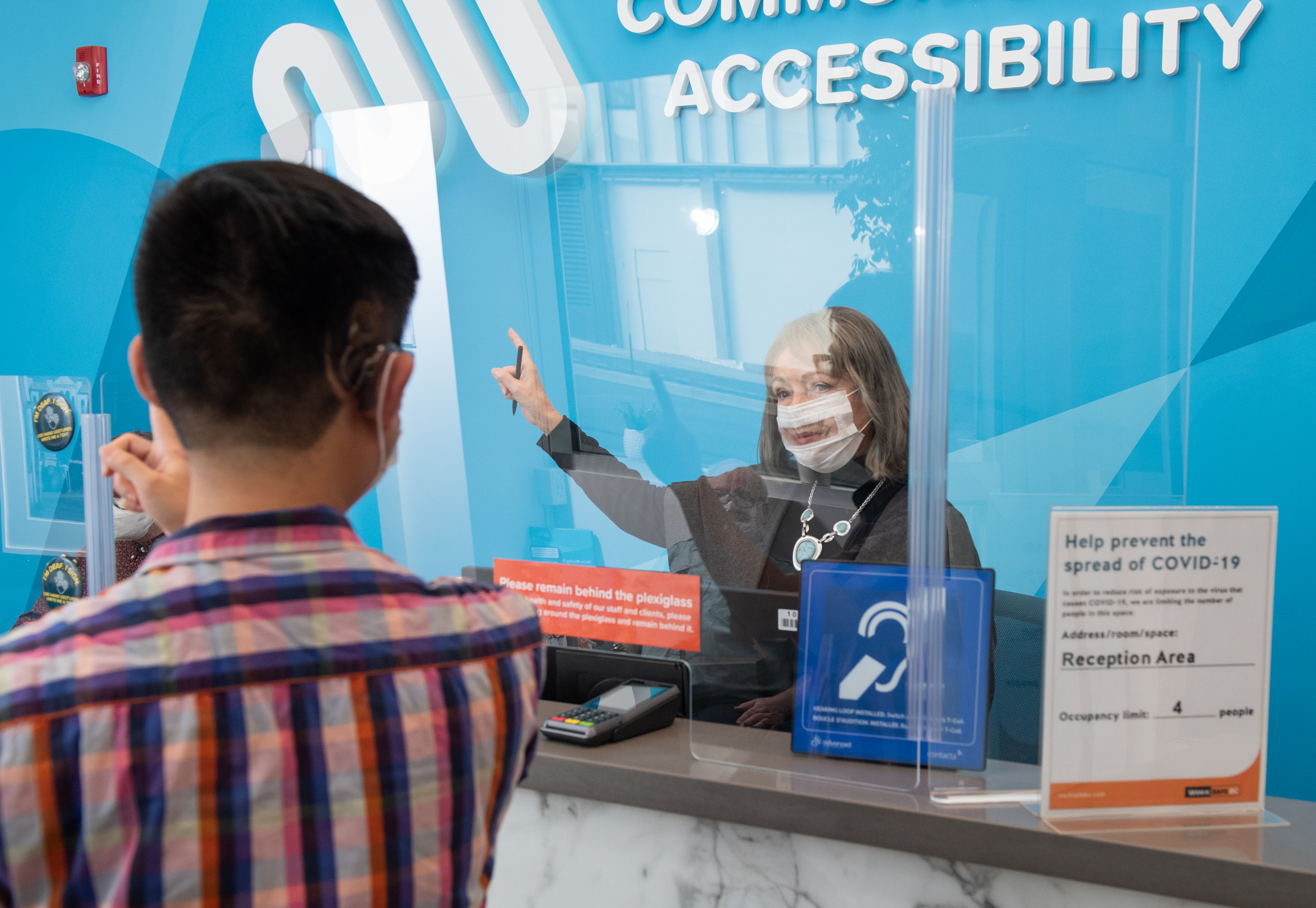 Man asking for information from a female receptionist behind plexiglass. She is wearing a clear face mask and is pointing beside her. There is a blue sign on the desk that has a white icon indicating the building is accessible for people with hearing loss.