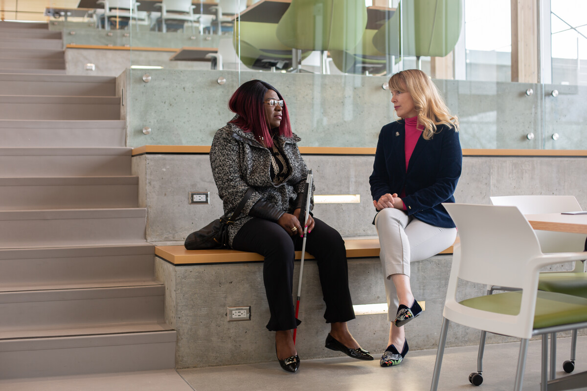 Two women sit in a modern indoor space engaged in conversation. One person uses a white cane.