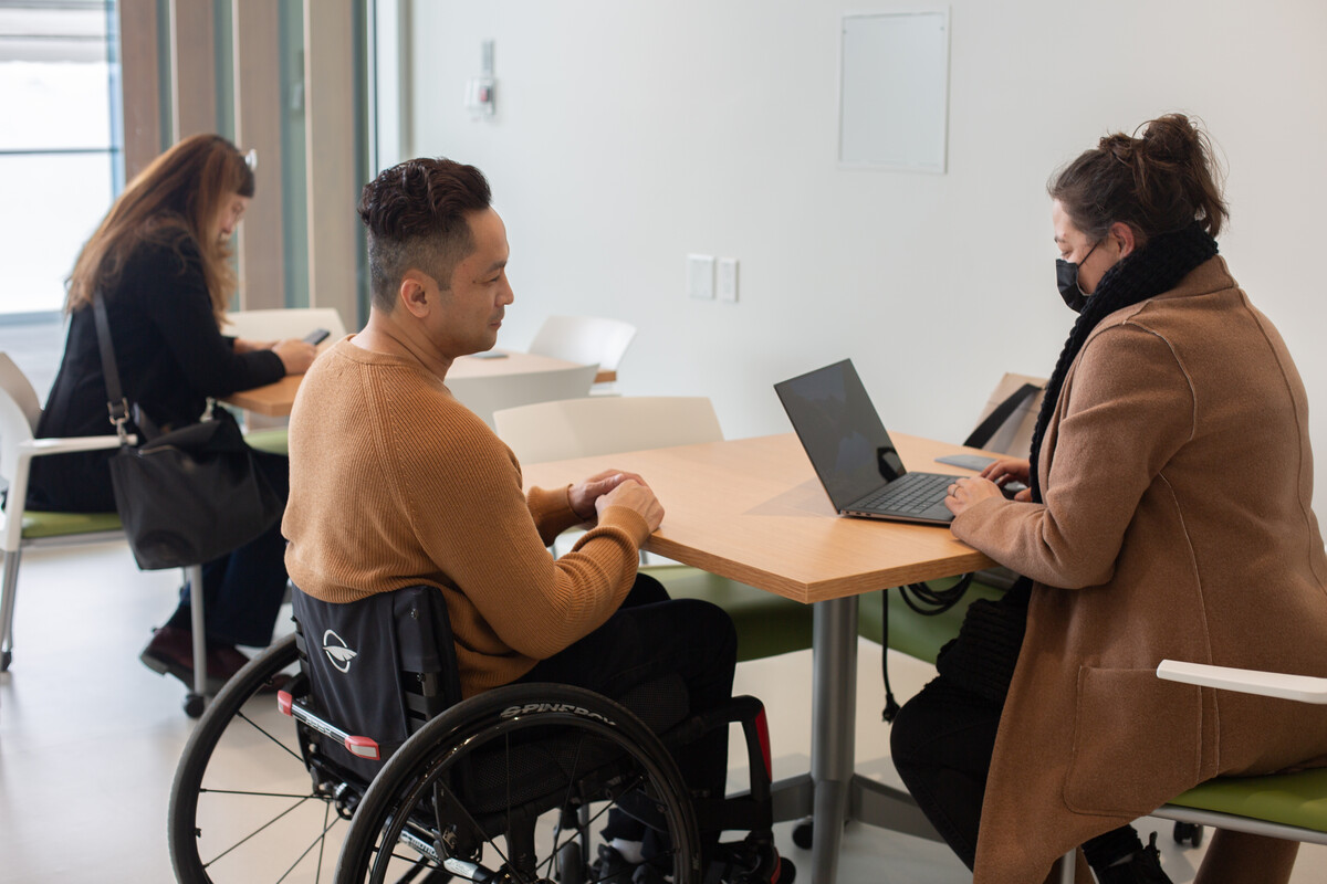 Two people at a table using a laptop. One person is using a wheelchair.