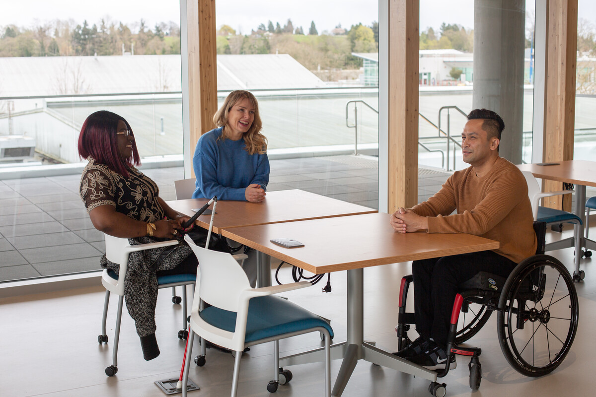 Three people of different ethnicities and abilities sitting at a table talking. One person has a white and red cane and other person is using a wheelchair.
