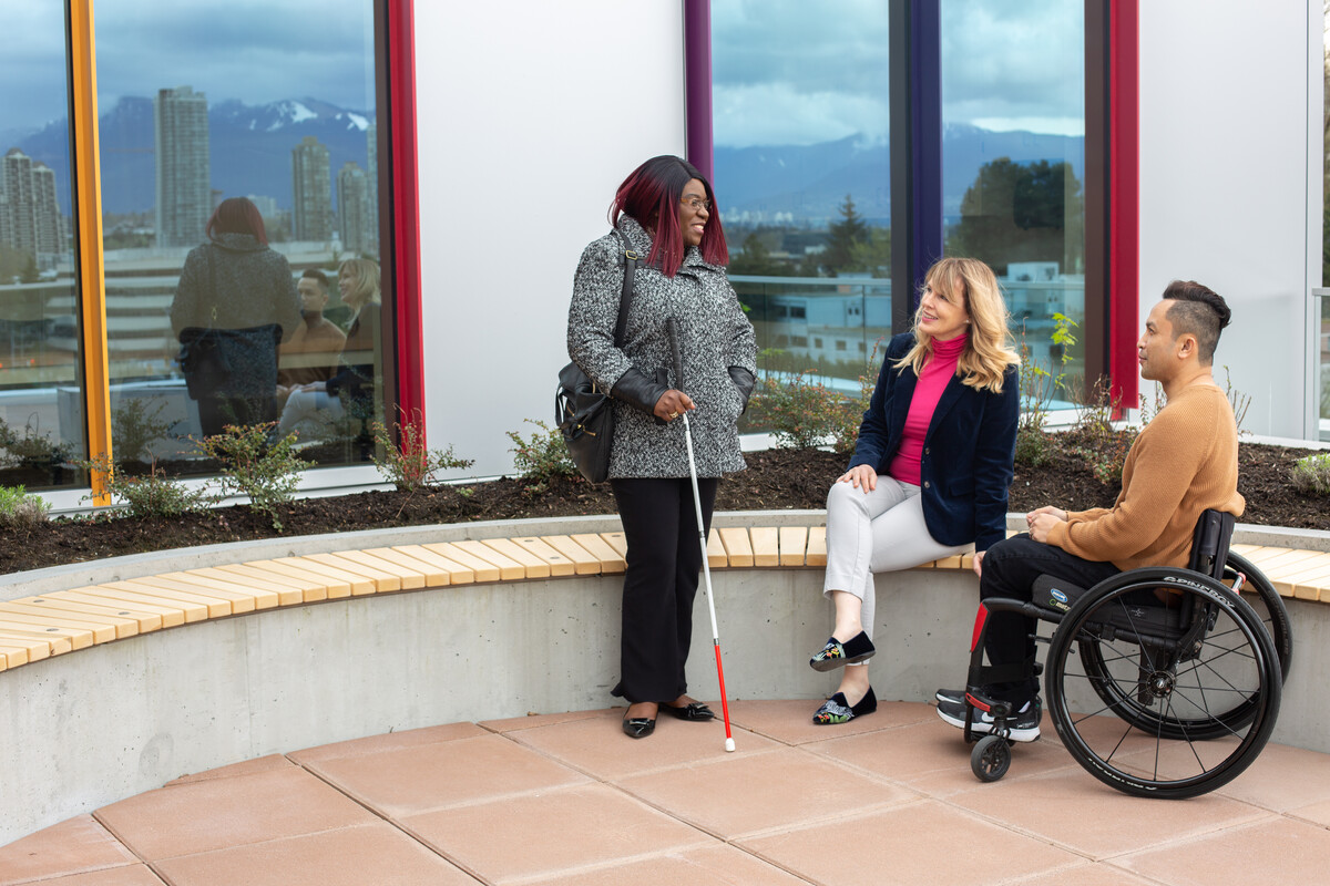 Three people outside talking next to a building. They each have a different disability 